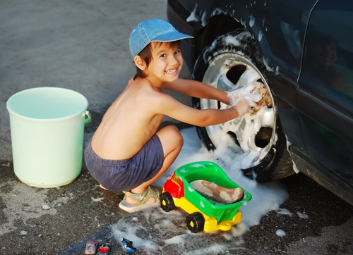 7 year old boy washing tires in the summer to with toy truck sponge and soapy suds splashing on pavement and side of car